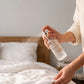 A person in a cream robe sprays a clear bottle of fragrance mist onto a neatly made white bed in a minimalist bedroom. The bottle label reads 'Scent of Nature'.