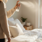 Person standing by a bed holding a CozyLife Clean Calm Linen Spray bottle in a bedroom setting.